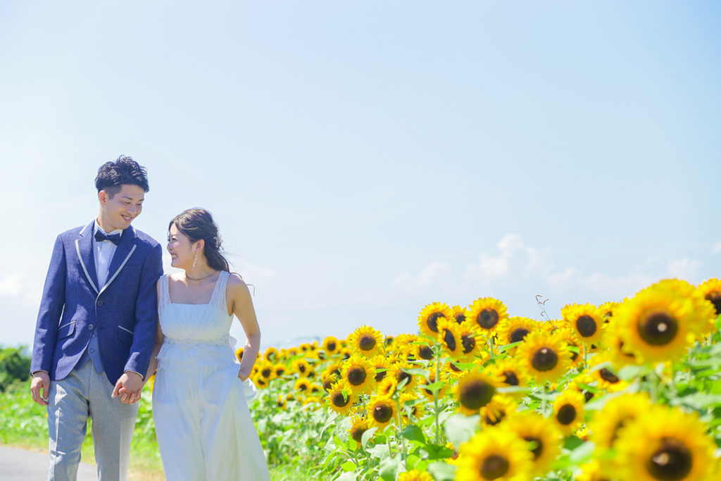 ひまわり畑の前を手をつないで笑顔で歩く新郎新婦