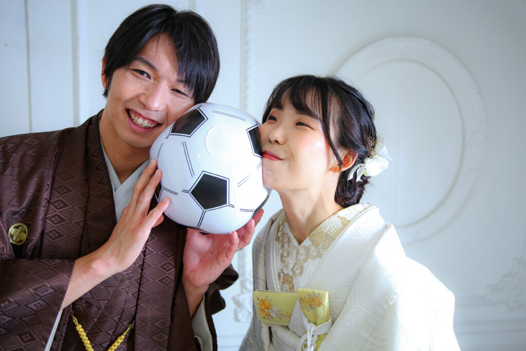 ほっぺたにサッカーボール風のボールを挟んだ笑顔の新郎新婦