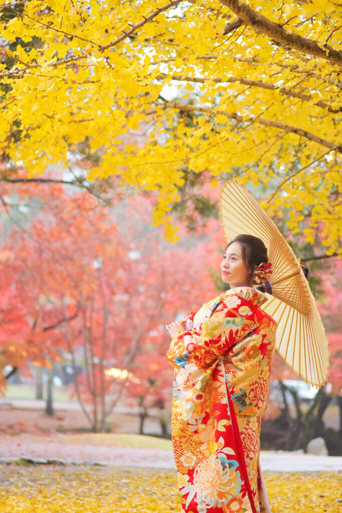 赤や黄色の紅葉黄葉の下で、和傘で佇む和装の新婦