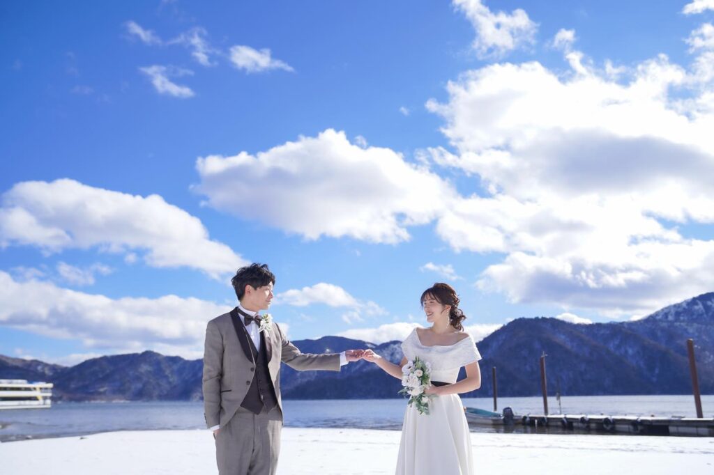 青空の下、雪の中禅寺湖畔で見つめあうドレス&タキシードのおふたり。
