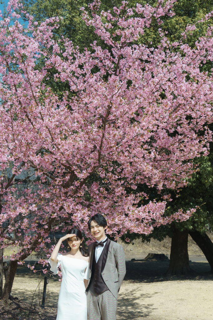 桜の木の下で、陽射しをまぶしそうに手で遮るドレスの花嫁様、隣で笑顔の花婿様。