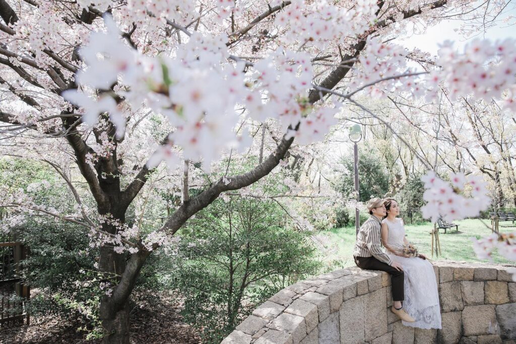 桜の木の下で、ナチュラルな衣装の新郎新婦が石塀の上に腰かけている。